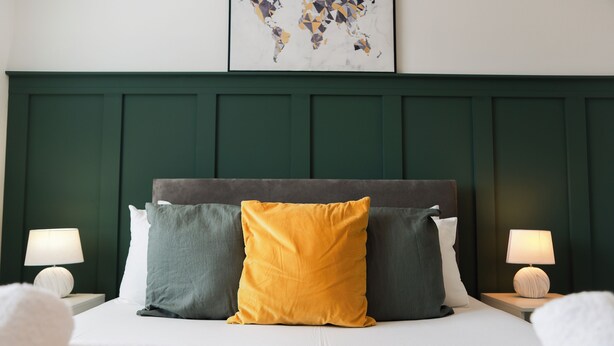 Una cama con cabecera verde, tres almohadas de distintos colores y dos lámparas de noche.