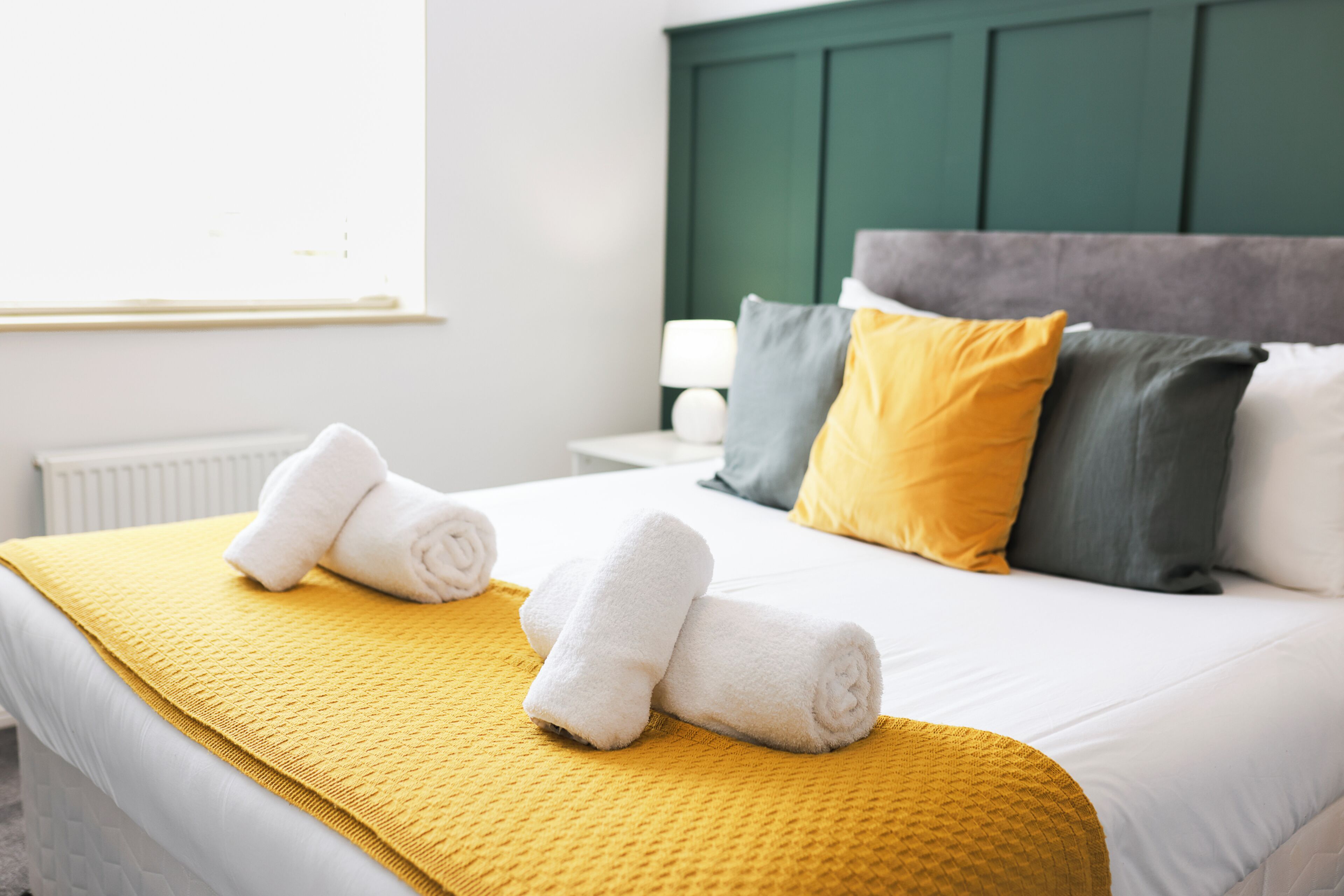 Un lit bien fait, avec du linge de lit blanc et un plaid jaune, une tête de lit verte et une table de chevet avec une lampe.