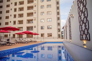 Ein Hotelpoolbereich mit Liegestühlen und roten Sonnenschirmen, umgeben von einem hohen Gebäude mit Balkonen.