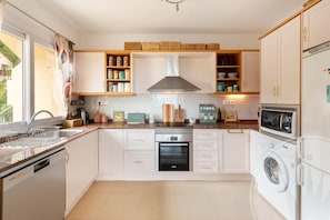 A modern kitchen with white cabinetry, a built-in oven, a dishwasher, a washing machine, and a window with a view of greenery. - La Manga Club Monte Verde Apartment 618 (Cartagena)