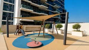 A playground with a slide, swings, and a circular play area, adjacent to a modern building with balconies. - Pure Voyage Damac Hills (Dubai)