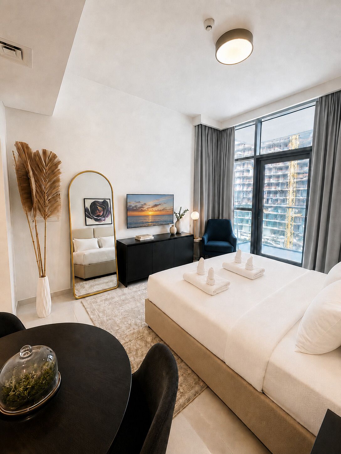 A modern hotel room with a large bed, a dining table, a TV, and a view of the city.