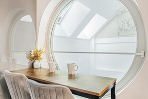 Ein Zimmer mit rundem Fenster, einem Holztisch, zwei Stühlen und einer Vase mit Blumen.