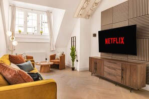 A living room with a sofa, armchair, coffee table, and a TV mounted on the wall displaying 'NETFLIX'. - Market Square Luxury | Modern 2-Bed City Retreat (Nottingham)