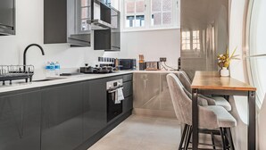 A modern kitchen with dark cabinetry, a stainless steel range hood, and a dining area with a wooden table and bar stools. - Market Square Luxury | Modern 2-Bed City Retreat (Nottingham)
