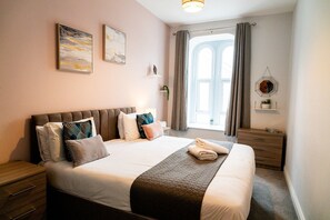 A neatly made bed with pillows, a bedside table, a nightstand, a window with curtains, and wall art. - Cosy Converted Chapel Retreat · Central Swansea (Swansea)
