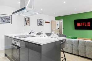 Una cocina moderna con una isla central, un área de comedor con sillas, un sofá y un televisor que muestra 'NETFLIX' en la pared.