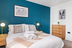 Ein ordentlich bezogenes Bett mit karierter Decke, zwei Nachttischlampen und eine Holzkommode mit Schubladen.