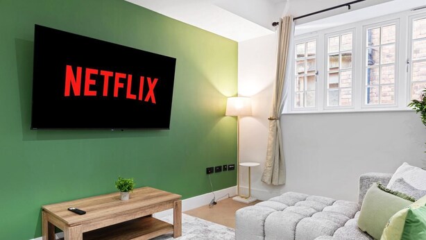 En stue med et fladskærms TV monteret på en grøn væg, et træsofabord, en grå sofa og en gulvlampe.