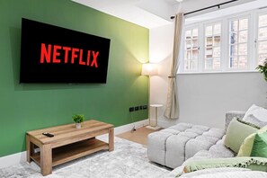Ein Wohnzimmer mit flachbildfernseher an einer grünen Wand, einem Holztisch, einem grauen Sofa und einer Stehlampe.