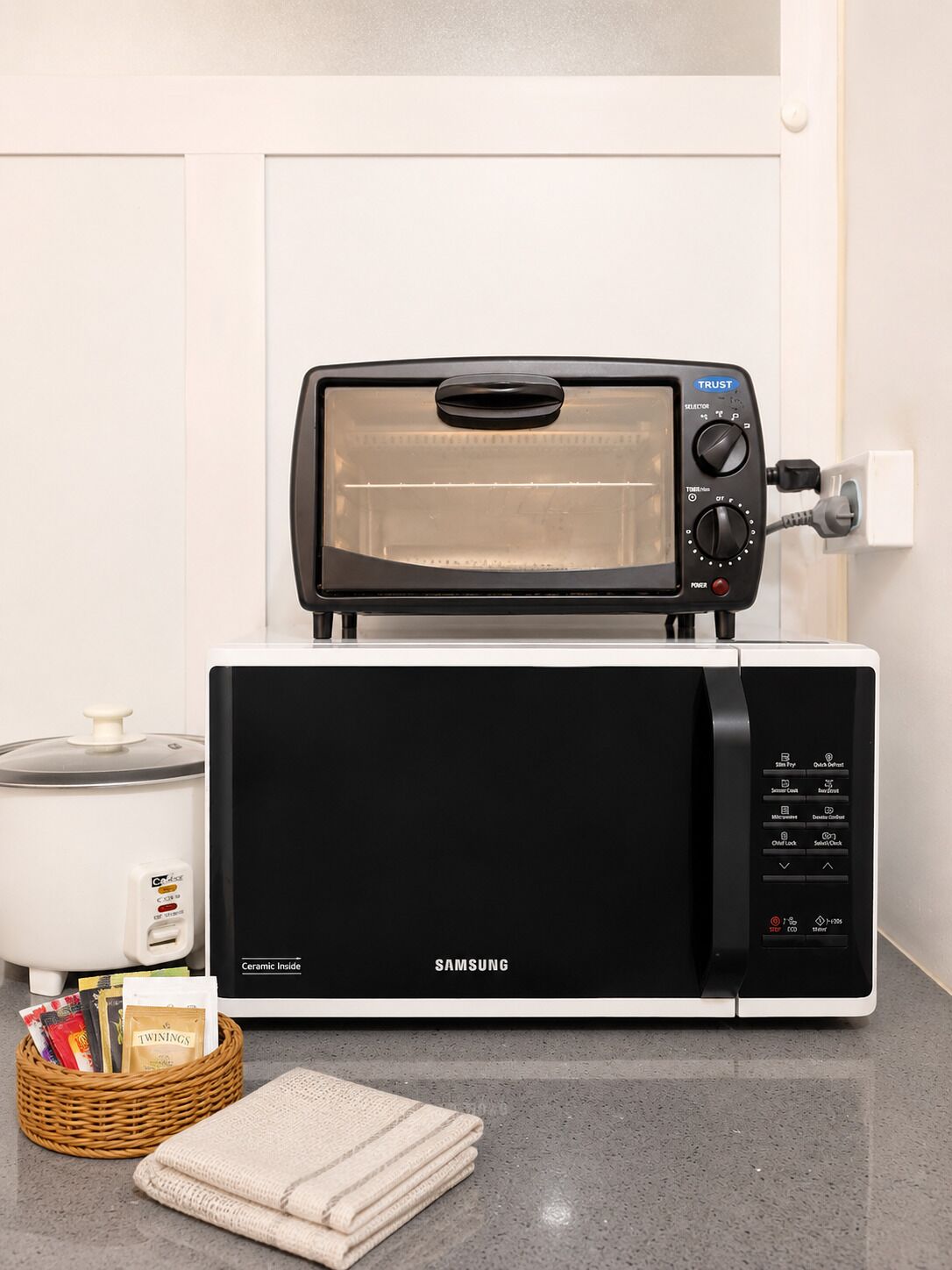 Un forno a microonde Samsung con un tostapane sopra, una pentola in ceramica e un cesto con oggetti su un piano di lavoro in cucina.
