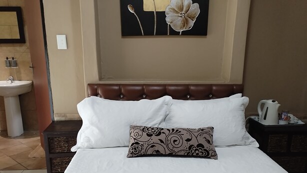 A neatly made bed with white linens and a decorative pillow, a brown headboard, a wall-mounted painting, and a bedside table with a kettle and cups.