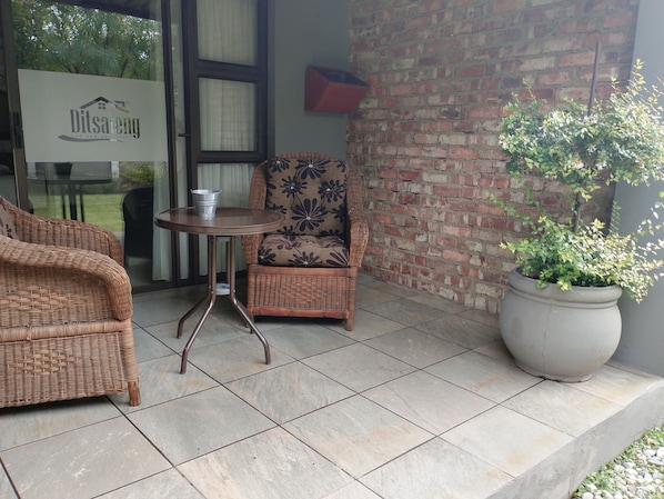 A wicker sofa and armchair with a patterned cushion, a small round table with a metal bucket, and a large potted plant on a tiled patio.