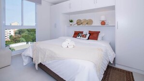 Un dormitorio moderno con un ventanal grande, una cama con ropa de cama blanca, una mesita de noche y vistas a edificios y árboles.