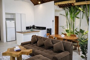 Un espace de vie moderne comprenant un canapé, une table basse en bois, une table à manger, un coin cuisine et de grandes plantes vertes.