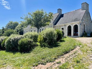 A stone building with a sloped roof, arched doorways, and a pathway leading to the entrance. - Soleil Levant Exceptional Villa with Pool (Quettehou)