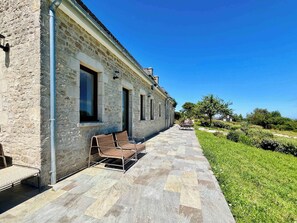 A stone building with a tiled patio, outdoor seating, and a clear blue sky. - Soleil Levant Exceptional Villa with Pool (Quettehou)