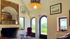 A room with a fireplace, leather sofas, a round table, a red chair, and large windows with views of greenery and the sea. - Soleil Levant Exceptional Villa with Pool (Quettehou)