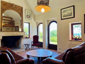 A room with a fireplace, leather sofas, a round table, a red chair, and large windows with views of greenery and the sea. - Soleil Levant Exceptional Villa with Pool (Quettehou)