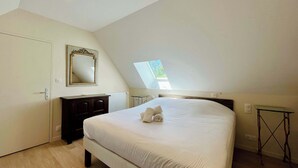 A bedroom with a bed, a dresser, a mirror, a skylight, and a small table. - Soleil Levant Exceptional Villa with Pool (Quettehou)