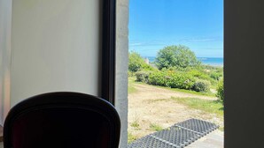 A view from inside a room with a window overlooking a grassy area and a clear blue sky. - Soleil Levant Exceptional Villa with Pool (Quettehou)