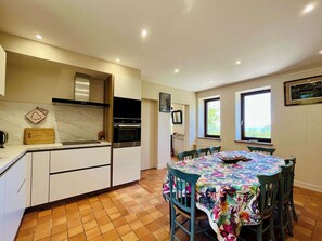 A modern kitchen with built-in appliances, a dining area with a floral tablecloth, and a view through the window. - Soleil Levant Exceptional Villa with Pool (Quettehou)