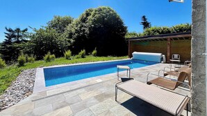 A swimming pool with lounge chairs and a covered area, surrounded by greenery. - Soleil Levant Exceptional Villa with Pool (Quettehou)