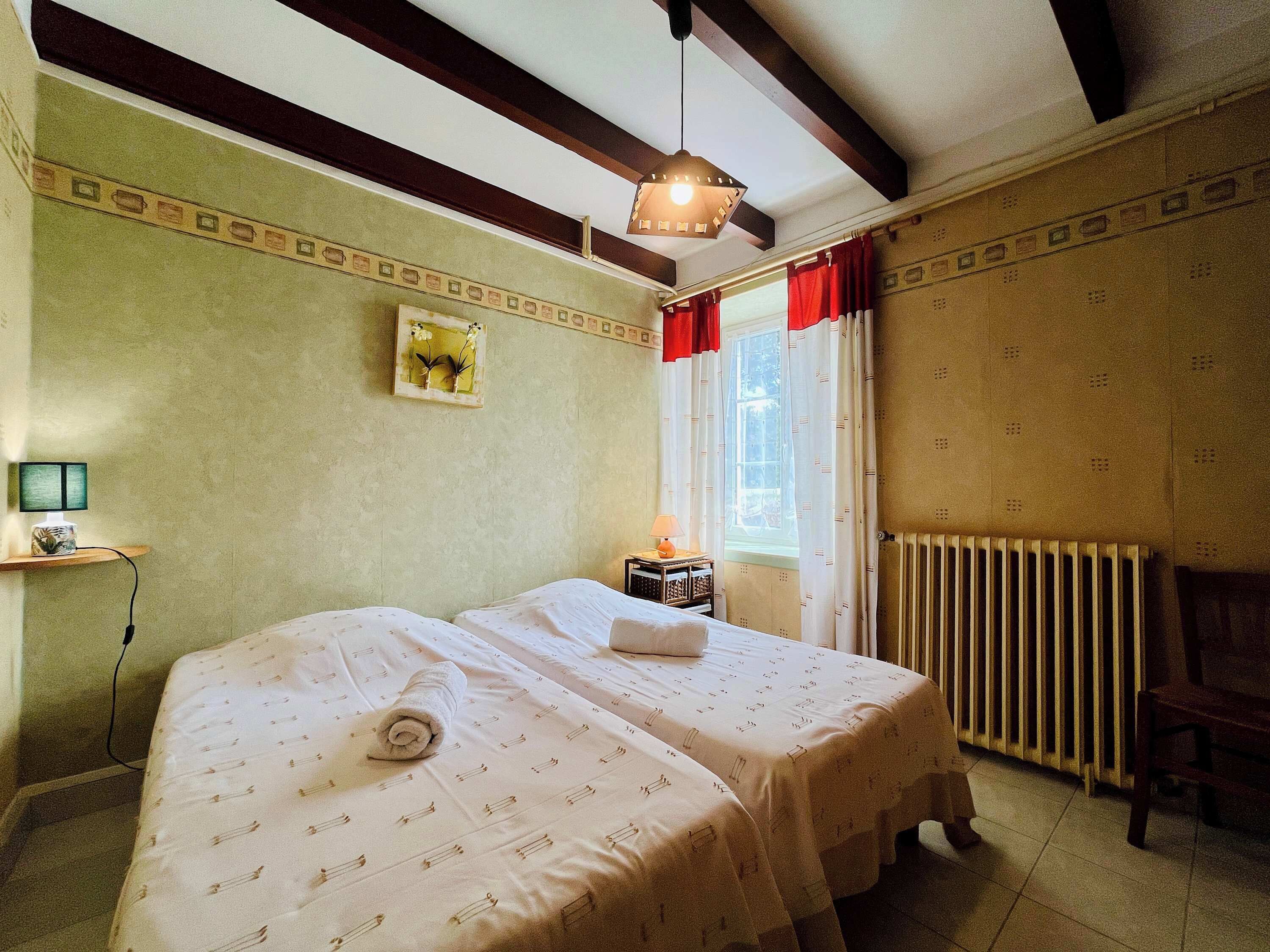A room with two beds, a radiator, a window with curtains, and a hanging light fixture.