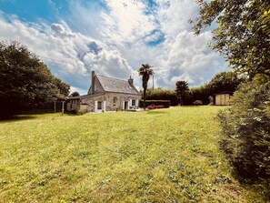 A stone house with a sloped roof, surrounded by a grassy yard and dense foliage. - Les Palmiers du Cotentin - 1 km from the sea (Aumeville-Lestre)