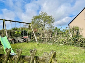 Ein Spielplatz mit Schaukeln und Rutsche, umgeben von einem Holenzaun und viel Grün.