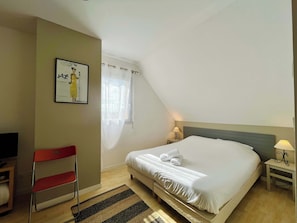 Ein Schlafzimmer mit Bett, Stuhl, Nachttisch, Lampe, Fenster mit Vorhängen und einem gerahmten Bild an der Wand.