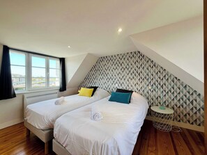 A bedroom with two beds, a window, a patterned wall, and a small round table. - La Maison Bleue, Authentic Fishermans House (Isigny-sur-Mer)
