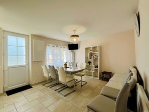 A modern living room with a sofa, a dining table with chairs, a bookshelf, and a television mounted on the wall.