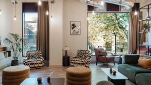 A modern living room with a sofa, armchairs, ottomans, a coffee table, and a lamp. There is a large window with curtains, a potted plant, and a framed picture on the wall. - Student Only YourTRIBE EC (London)