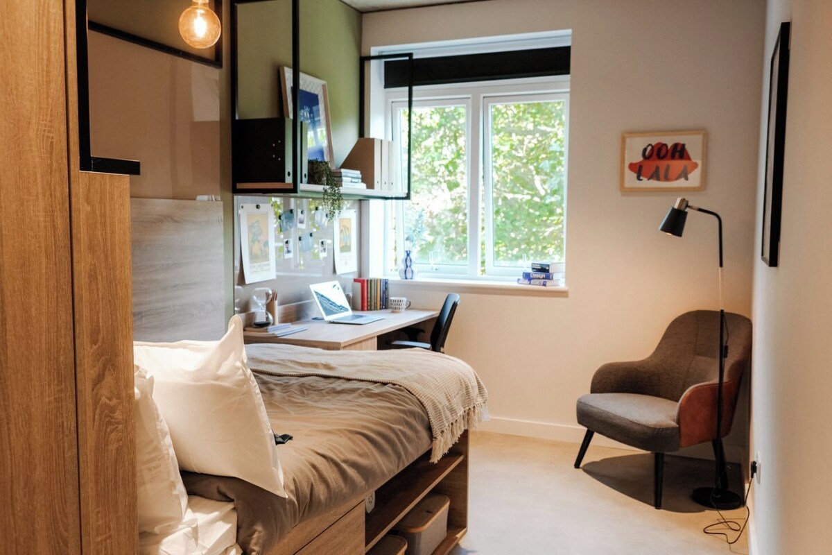 Ein Schlafzimmer mit Bett, Schreibtisch, Stuhl, Fenster und einem an der Wand befestigten Regal.