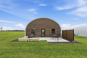 A modern building with a curved roof, a patio area with outdoor seating, and a greenhouse structure in the background.