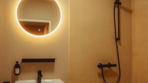 Uma casa de banho com um espelho redondo, uma pia, chuveiro e um porta-toalhas.
