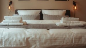 Uma cama com lençóis brancos, cabeceira marrom e duas luminárias fixadas na parede.
