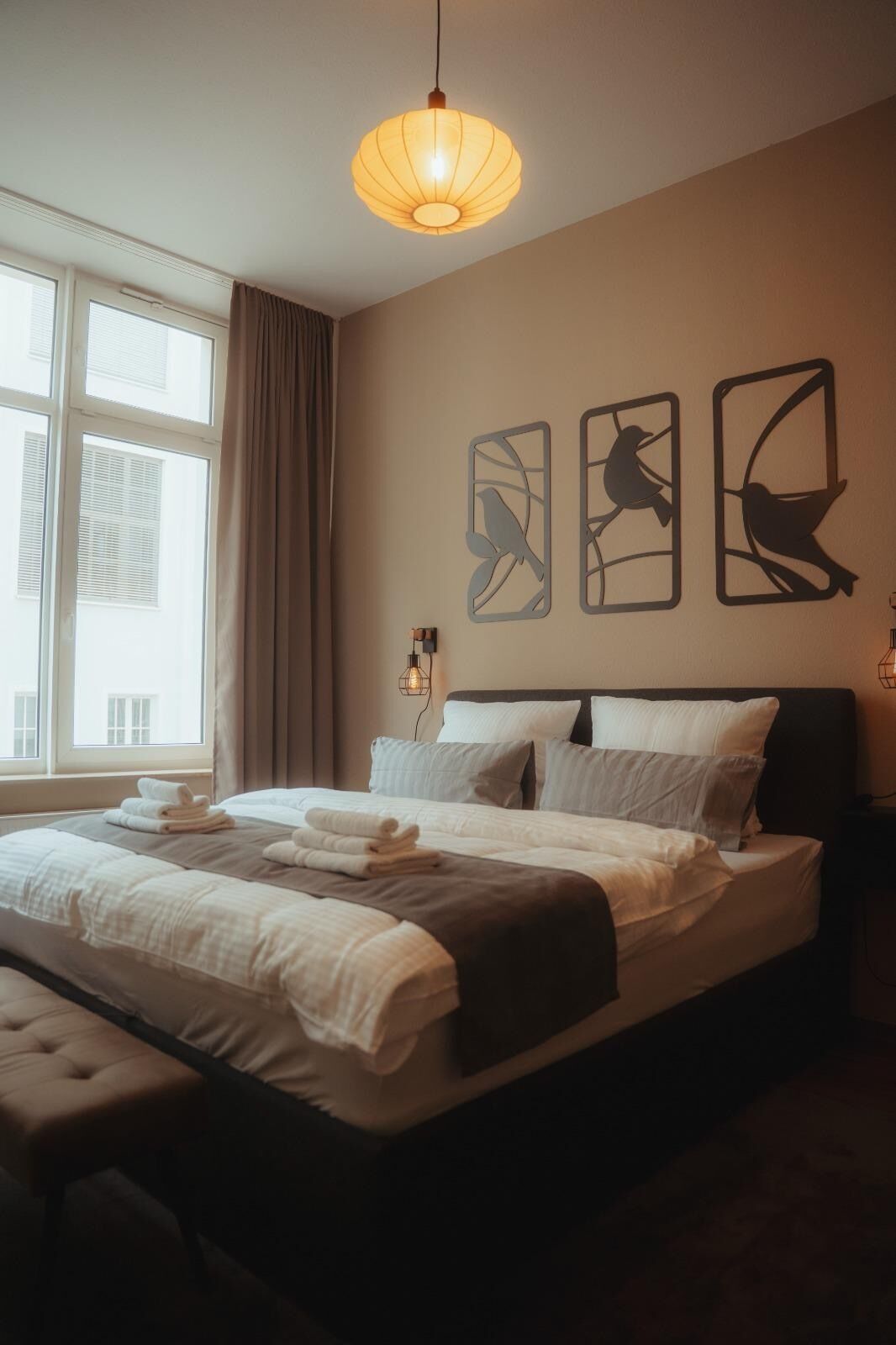 Ein ordentlich bezogenes Bett mit Kissen und Decke, eine Nachttischlampe und Vogelbilder an der Wand.
