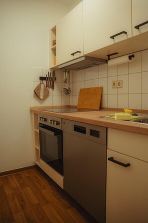 A kitchen with wooden cabinets, a built-in oven, a dishwasher, and a countertop with a cutting board and a sponge. - City Apartment I Free Parking (Bielefeld)