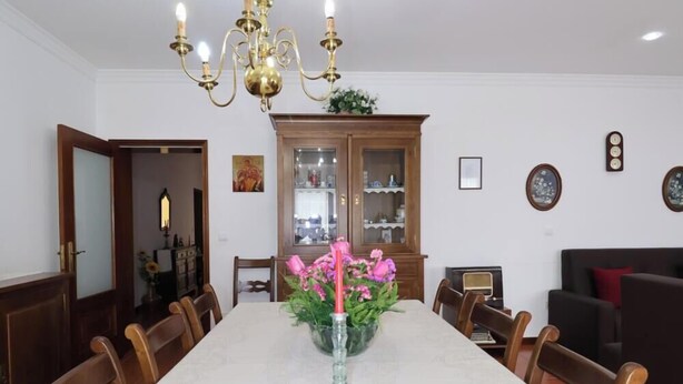 Ein Esszimmer mit einem langen Tisch, Stühlen, einer Chandelüster, einem Schrank mit Glastüren und einer Vase mit Blumen.