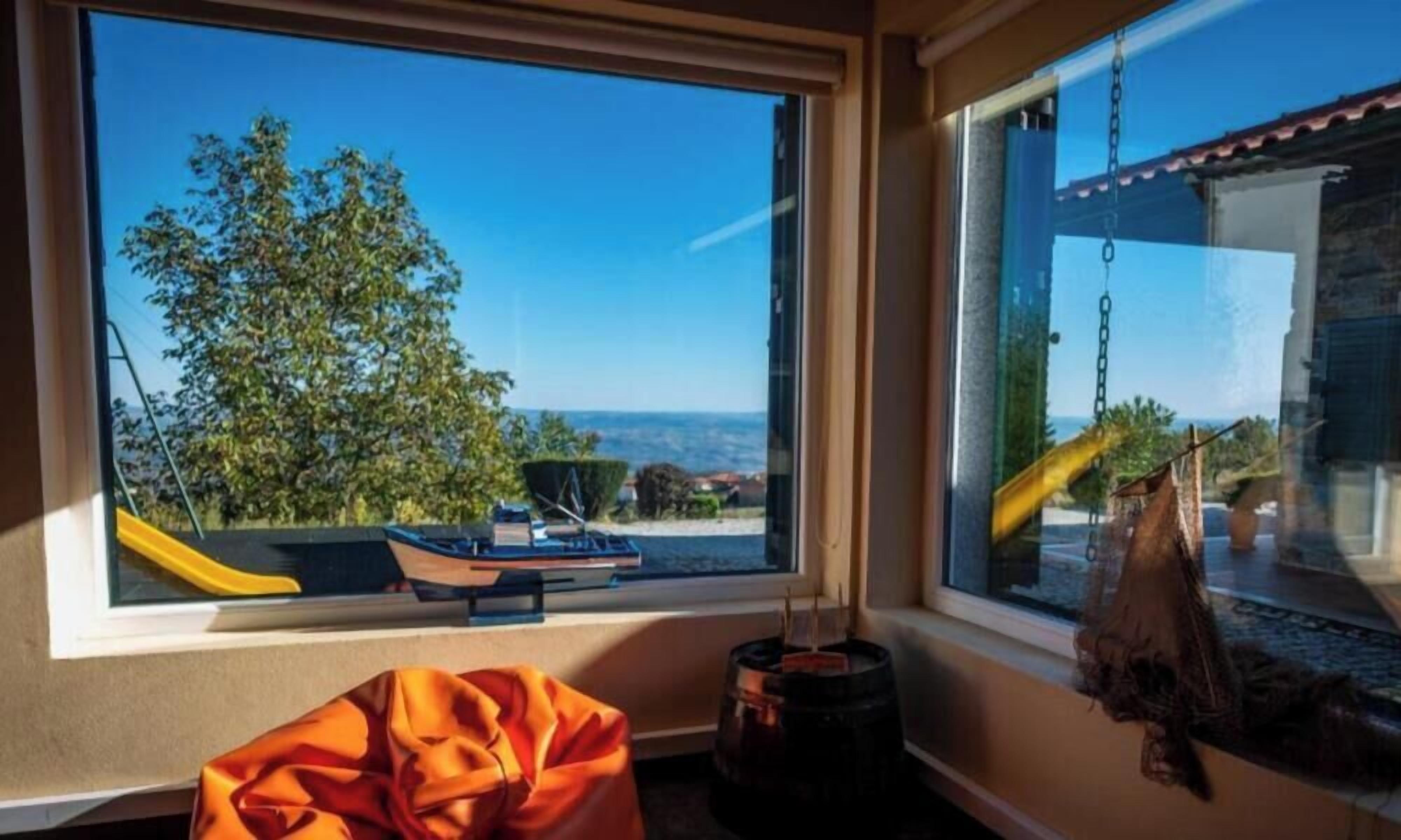 Ein Zimmer mit großem Fenster, das auf einen Spielplatz und einen Baum blickt, ein kleiner Tisch mit Stuhl und eine hängende Pflanze.