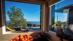 Ein Zimmer mit großem Fenster, das auf einen Spielplatz und einen Baum blickt, ein kleiner Tisch mit Stuhl und eine hängende Pflanze.
