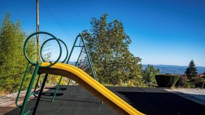 Ein Spielplatz mit Rutsche und Schaukeln, umgeben von Bäumen und blauem Himmel.