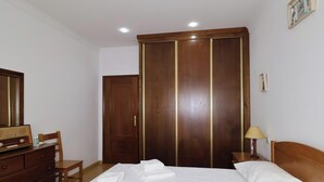 A hotel room with a bed, a dresser, a desk, a chair, a wardrobe, and a bedside table with a lamp.