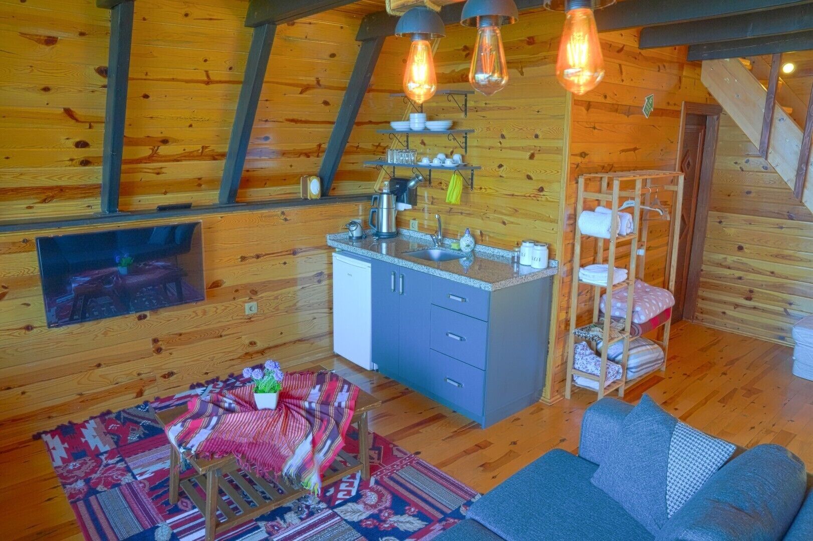A cozy cabin interior with a kitchenette, a sofa, a patterned rug, and a wooden shelf with towels.