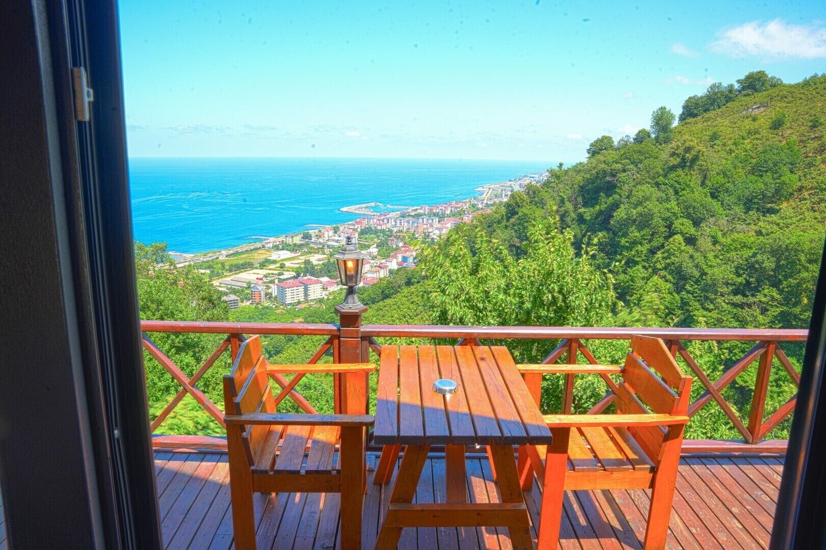 A balcony with a wooden table and chairs, overlooking a coastal town and lush greenery.