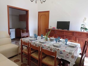 A dining area with a table set for four, surrounded by wooden chairs with woven seats. A television is mounted on a wooden cabinet above the table. - Salento Vacation Home "Nonno Totò" with Garden, Stone Barbecue, and Outdoor Shower (TAVIANO)