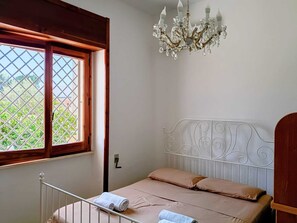 A bedroom with a metal bed frame, a window with a wooden frame, and a chandelier. - Salento Vacation Home "Nonno Totò" with Garden, Stone Barbecue, and Outdoor Shower (TAVIANO)