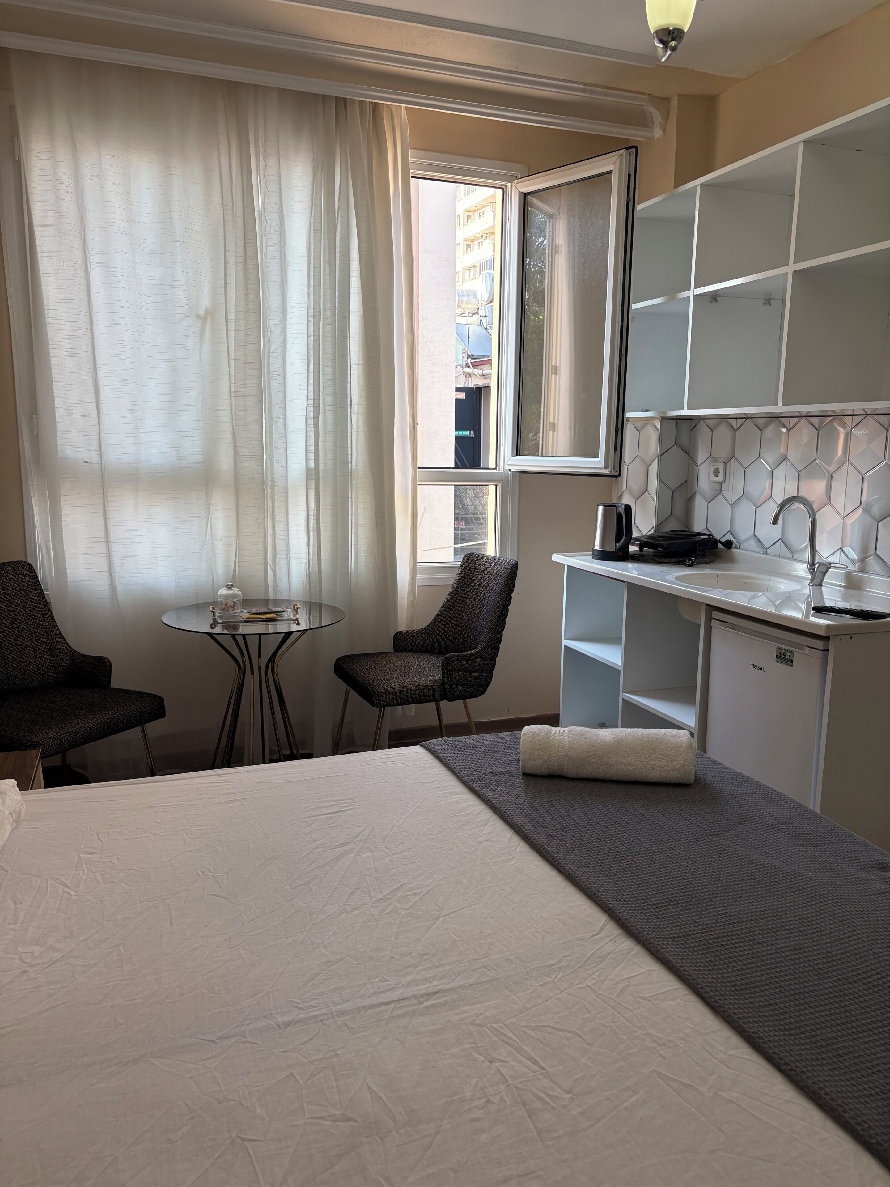A hotel room with a bed, a small table, two chairs, a kitchenette, and a window with curtains.